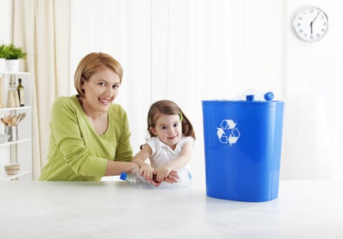 A woman with short blonde hair wearing a light green top is seated at a white table, smiling at the camera while a young girl with long brown hair in pigtails, dressed in a white shirt, sits beside her. In front of them on the table is a clear plastic water bottle. To their right, there is a large blue recycling bin with a white recycling symbol on its front, positioned on a clean, light-colored surface. The background features a bright, modern kitchen with a white wall, a white clock, and some shelves with small decorative items. The scene appears well-lit with natural daylight, suggesting a domestic environment focused on waste sorting and recycling, consistent with rubbish and waste management themes often associated with commercial waste removal services in Bishops Stortford.