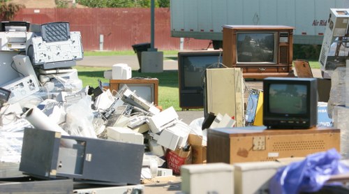 A large outdoor pile of discarded electronic devices and waste materials, including numerous old CRT televisions with wooden casings, monitors, and computer peripherals, stacked haphazardly on a paved surface. Some units are leaning against each other, with cables and plastic packaging visible among the clutter. The background shows a grassy area with a red fence and trees, indicating the waste collection is set in a suburban or industrial environment. The scene is illuminated by natural daylight, highlighting the variety of textures and colors, predominantly off-white, black, brown, and grey, associated with the electronic waste. The clutter appears ready for collection or disposal, illustrating a typical electronic and rubbish removal scenario handled by companies like Commercial Waste Bishops Stortford in the local area.