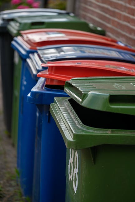 A row of large outdoor wheelie bins lined up along a pavement beside a brick wall, with the bins in various colours including green, blue, red, and dark green. The bins are made of durable plastic wit